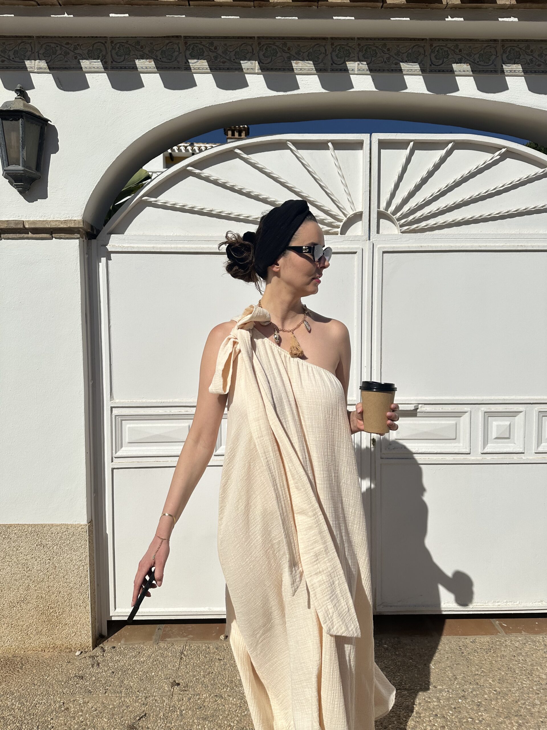 Modelka w kremowej sukience na lato z muślinowej bawełny, asymetrycznej na jedno ramię, z szarfą w talii – Kadora Studio.